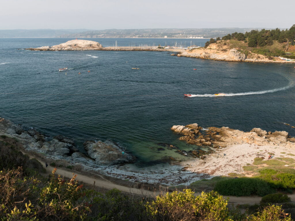 Vista panorámica de la bahía de Algarrobo con embarcaciones navegando entre kayaks y lanchas, un islote rocoso, una caleta de aguas turquesa con playa de arena blanca y un sendero costero en primer plano, parte del Litoral de los Poetas en la Región de Valparaíso.