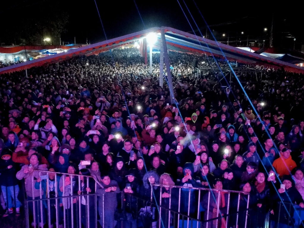 Miles de personas se reúnen bajo una gran carpa iluminada durante un concierto nocturno en una de las fondas patrias, con celulares en alto captando el momento.