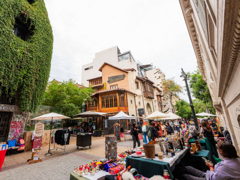 Calle del Barrio Lastarria en Santiago con puestos de artesanía y feria callejera, sombrillas blancas, fachadas de arquitectura patrimonial de madera y personas paseando entre edificios modernos y antiguos, uno de los barrios turísticos ideales para escapadas de fin de semana sin auto.