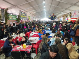Interior de una fonda techada con largas hileras de mesas ocupadas por comensales, entre banderines, guirnaldas y globos que decoran el ambiente dieciochero.