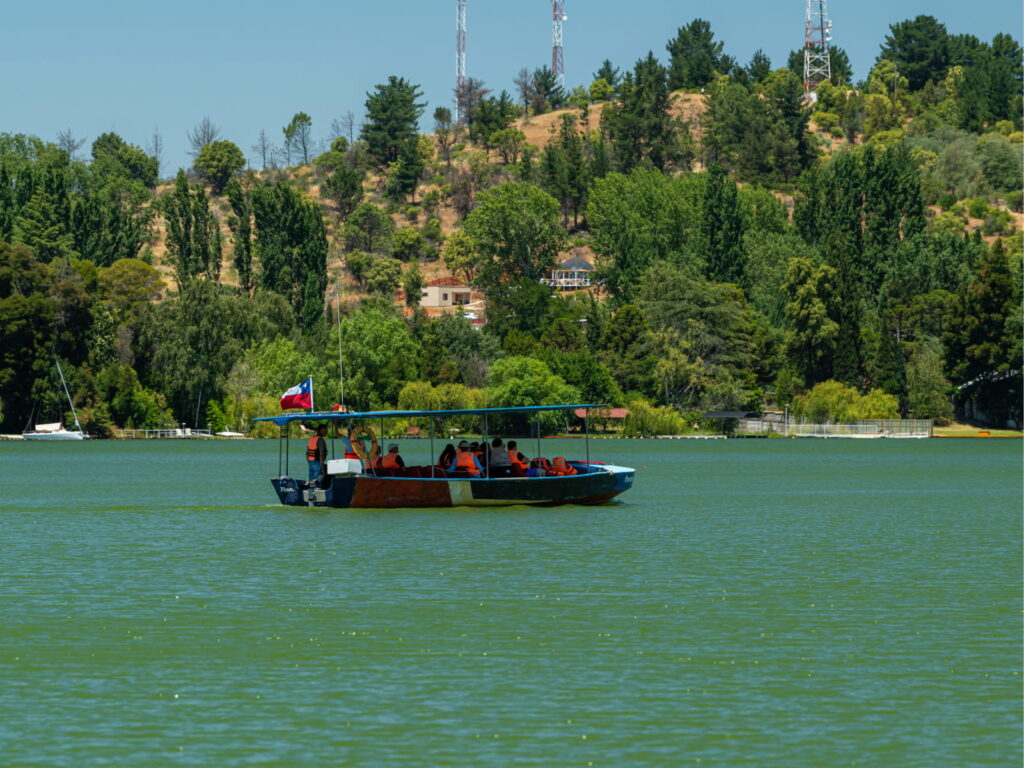 Bote turístico con pasajeros usando chalecos salvavidas navegando por la Laguna Avendaño en Quillón, rodeado de cerros boscosos, destino del Valle del Itata.