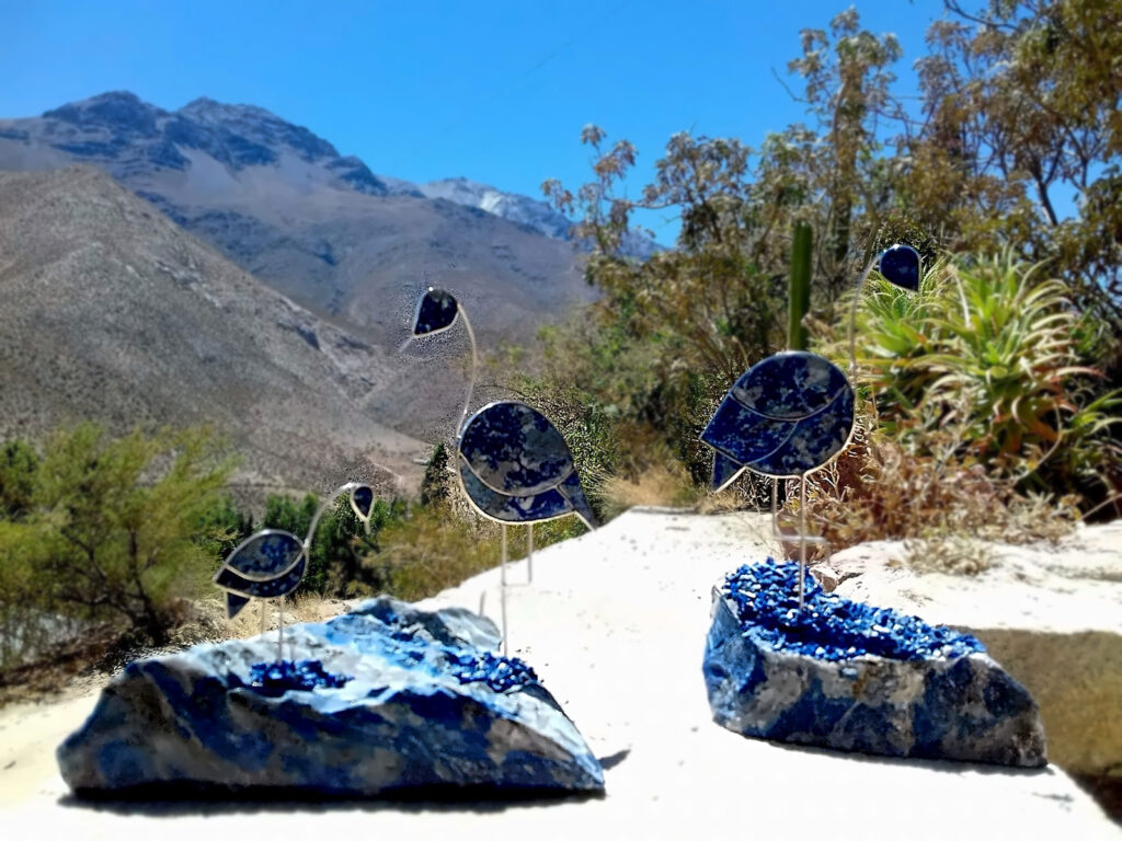 Piezas artesanales de lapislázuli, la piedra azul típica de Monte Patria, con forma de flamencos montados sobre rocas del mineral frente a un paisaje de cerros y vegetación del valle.