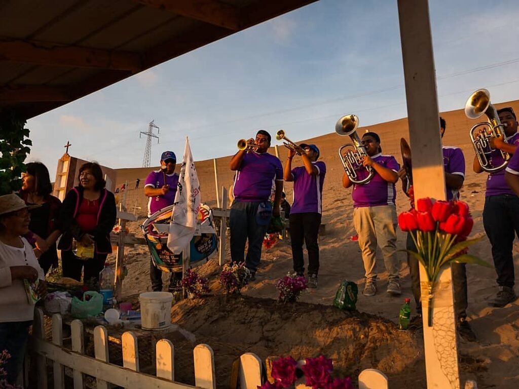 Banda de bronces con poleras moradas toca trompetas y bombardinos junto a una tumba en un cementerio de Arica, mientras mujeres de la familia escuchan al atardecer.