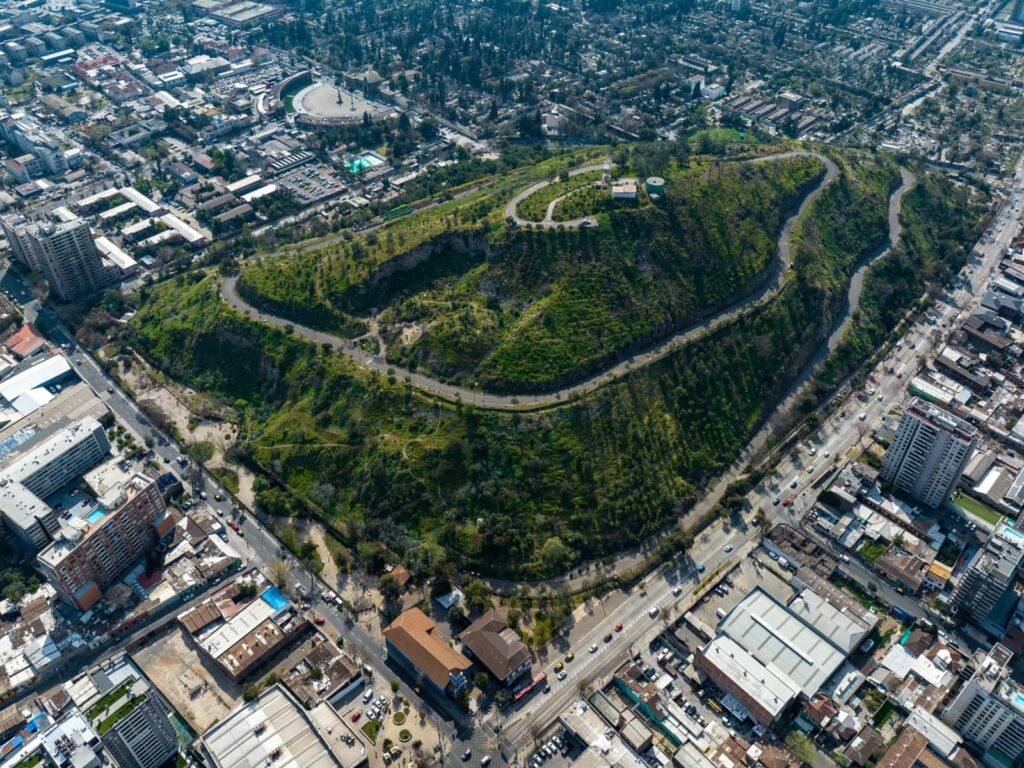 Vista aérea del Cerro Blanco, en Recoleta, con su camino sinuoso que rodea la cima entre laderas verdes y los edificios, calles y avenidas de la ciudad alrededor