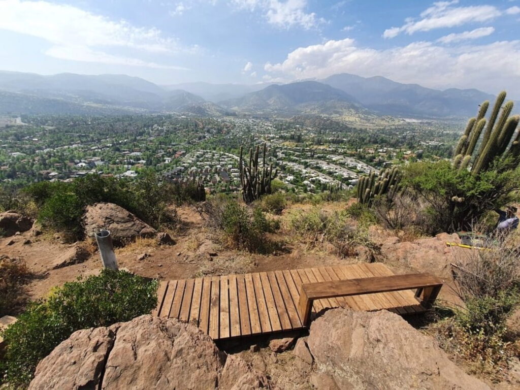 mirador de madera con banca entre rocas y quiscos en el Cerro del Medio, en Lo Barnechea, con vista al valle, las casas del sector y los cerros del fondo