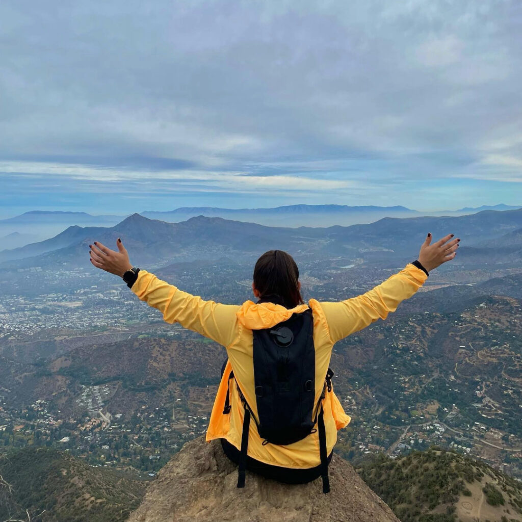 excursionista con chaqueta amarilla y mochila de hidratación, sentada con los brazos abiertos sobre una roca en la cumbre del Cerro Pochoco, en El Arrayán, con vista al valle y las montañas