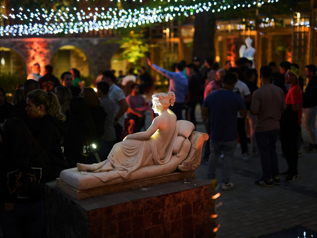 Escultura de mármol de una mujer recostada, iluminada en un patio con arcos y guirnaldas de luces, donde un grupo de personas comparte de noche en el Hotel Leonera.