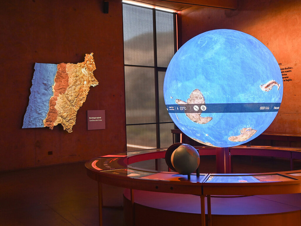 Sala de museo con un globo terráqueo interactivo iluminado que proyecta la Tierra en otra era geológica. Al fondo, en la pared, hay un mapa en relieve de la morfología regional con la Cordillera de la Costa.