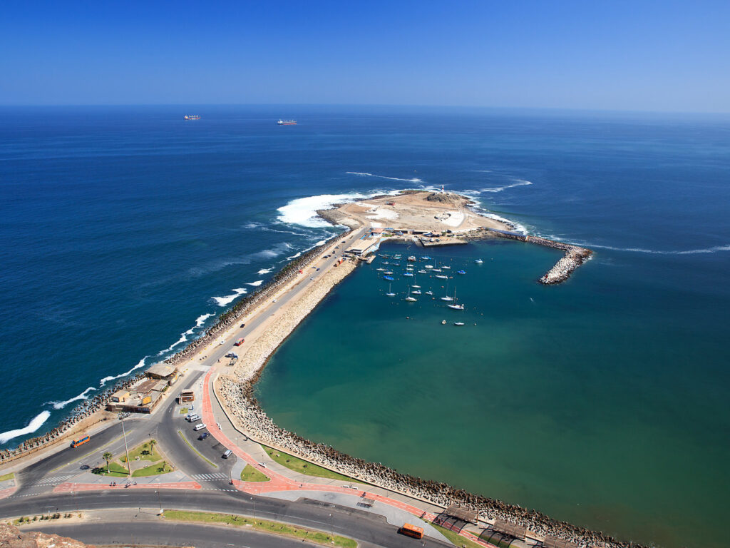 Vista aérea desde el Morro de Arica hacia la Isla del Alacrán, unida a la costa por un rompeolas. Se ven veleros anclados en la bahía, la costanera y el océano Pacífico de fondo, en la región de Arica y Parinacota.