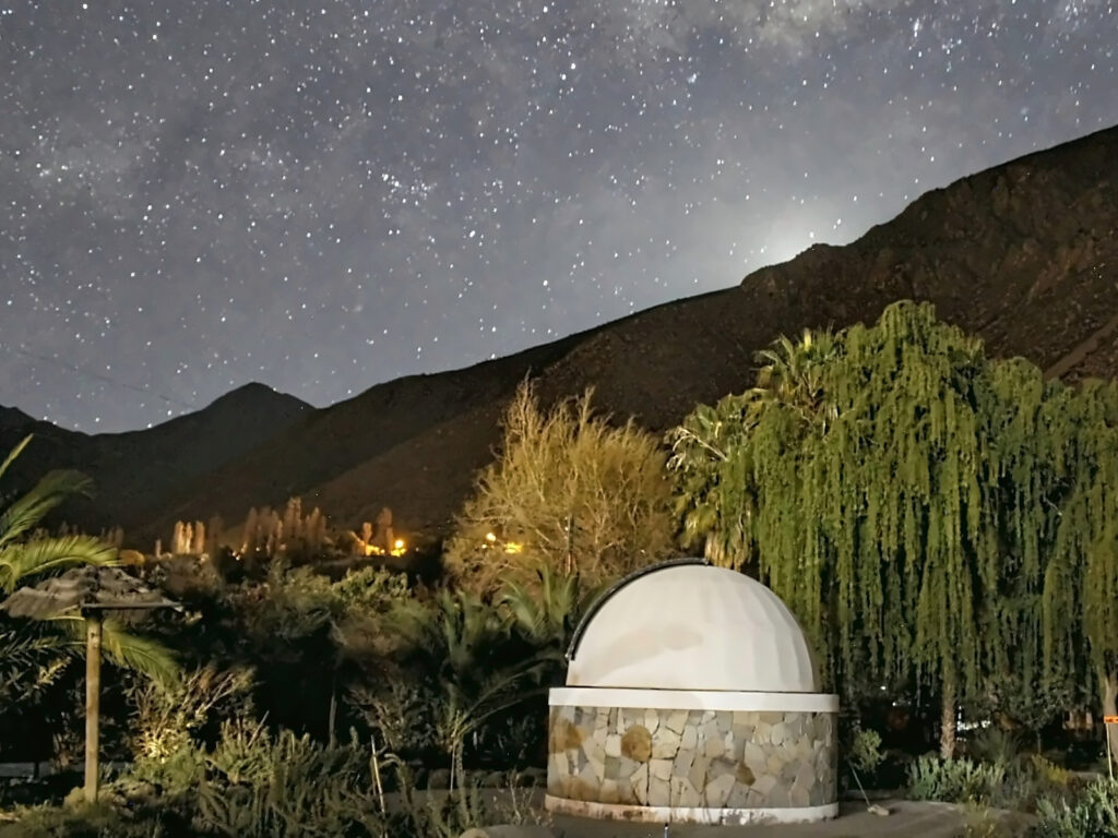Noche estrellada sobre el Observatorio Valle del Sol en Monte Patria, con la Vía Láctea sobre los cerros y las luces del pueblo entre los árboles del valle.