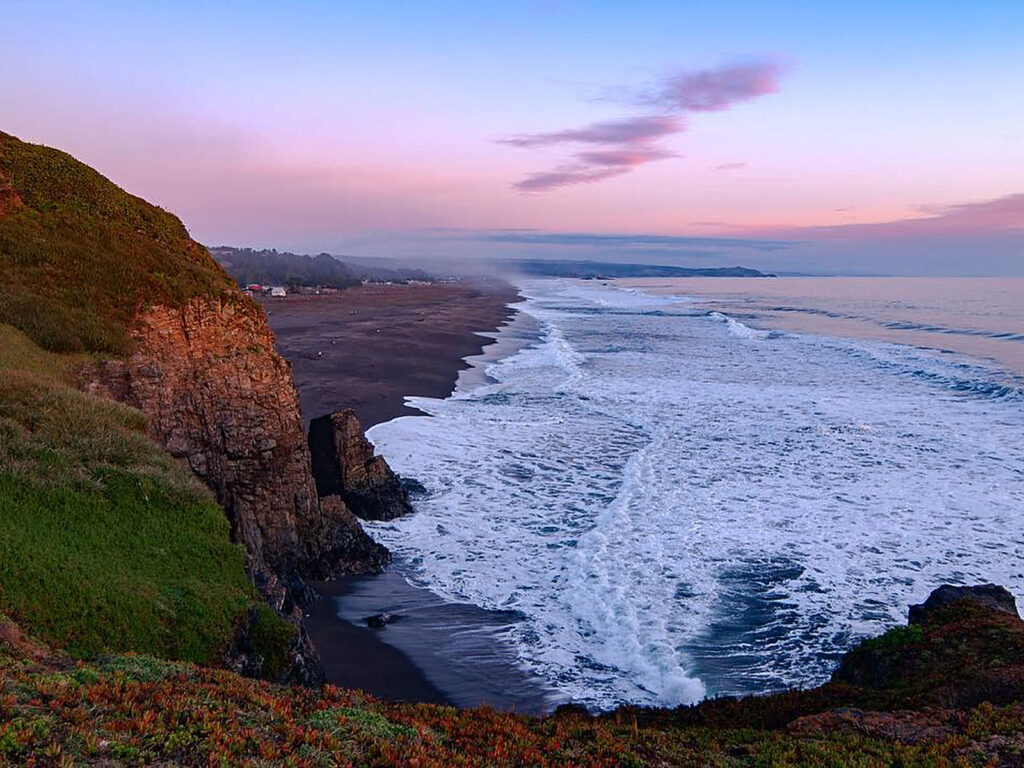  Acantilado cubierto de pasto y rocas frente a una extensa playa de arena negra con oleaje blanco, bajo un cielo rosado al atardecer en Buchupureo. 