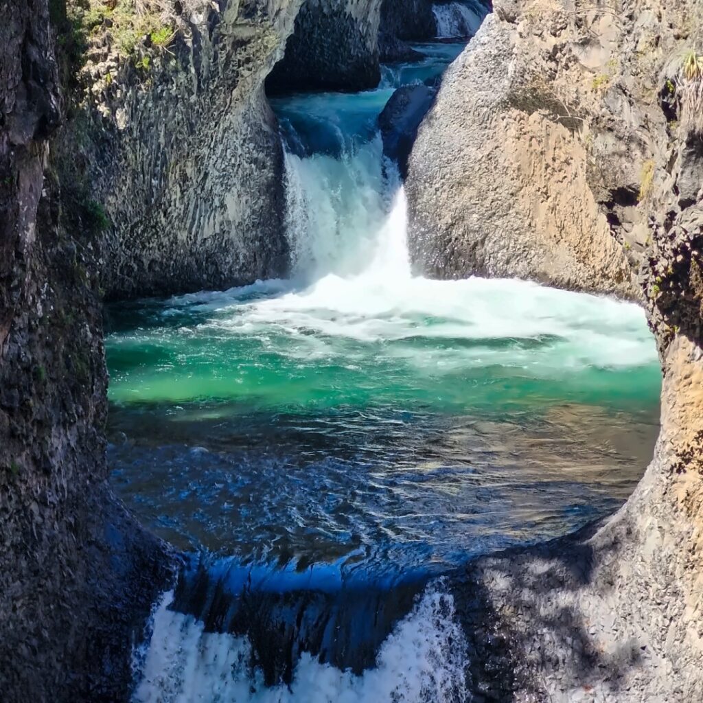 Cascada de aguas turquesa entre paredes de roca volcánica en el Parque Nacional Radal Siete Tazas, con una segunda caída de agua en primer plano.