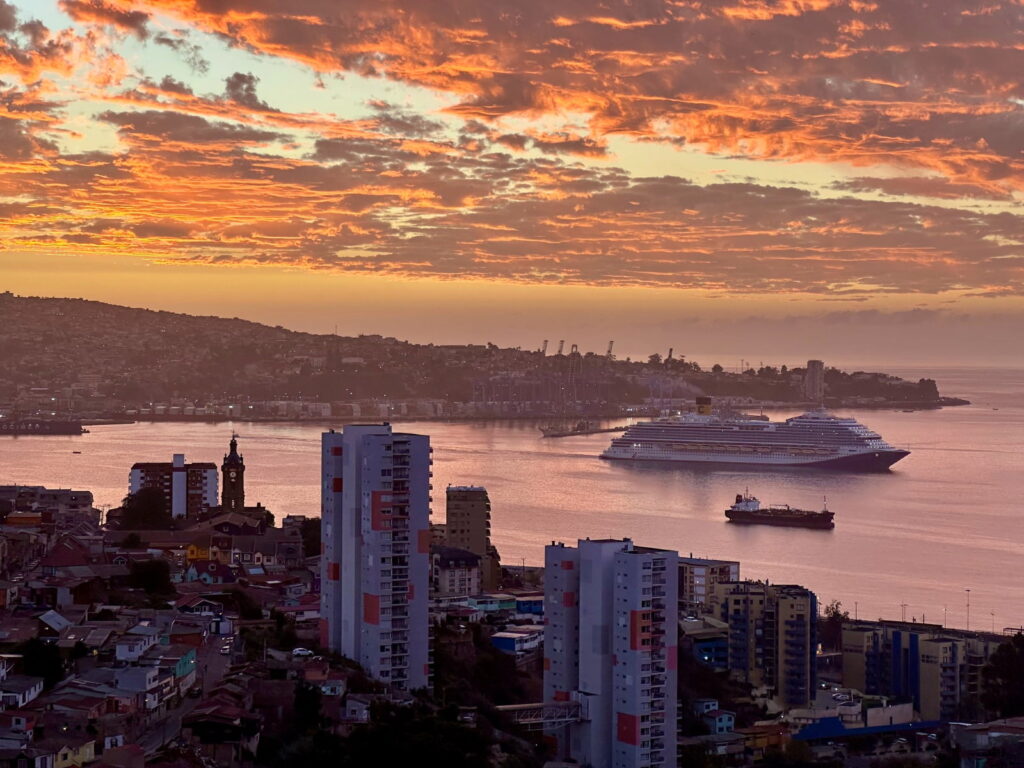 Crucero navegando por la bahía de Valparaíso al atardecer, con cerros de casas coloridas y cielo anaranjado, en Chile.